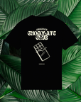 Un t-shirt noir avec un design au dos représentant une tablette de chocolat illustrée de façon minimaliste. Le texte 'Origines Chocolate Club' est inscrit en haut en lettres stylisées, suivi de l'inscription 'Bordeaux' en bas. Le t-shirt est posé sur un fond de grandes feuilles vertes naturelles.