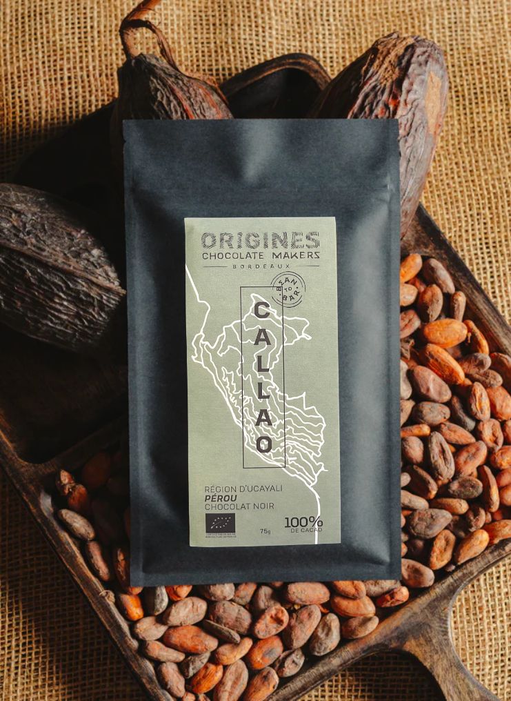 Tablette de chocolat noir Origines Chocolate Makers, emballée dans un sachet noir avec une étiquette verte indiquant 100 % de cacao provenant du Pérou, entourée de fèves de cacao et de cabosses sur un fond en toile de jute.
