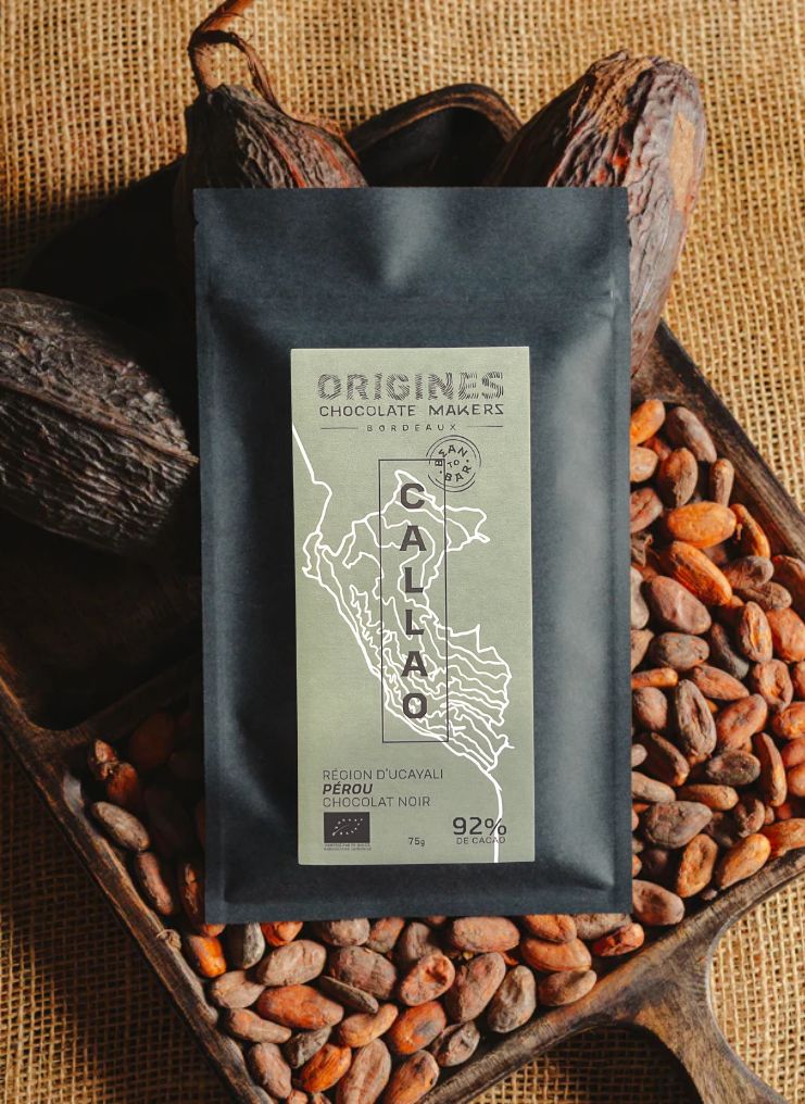 Tablette de chocolat noir Origines Chocolate Makers, emballée dans un sachet noir avec une étiquette verte indiquant 92 % de cacao provenant du Pérou, entourée de fèves de cacao et de cabosses sur un fond en toile de jute.