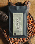 Tablette de chocolat noir Origines Chocolate Makers, emballée dans un sachet noir avec une étiquette verte indiquant 92 % de cacao provenant du Pérou, entourée de fèves de cacao et de cabosses sur un fond en toile de jute.