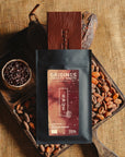 Tablette de chocolat noir au grué Origines Chocolate Makers, emballée dans un sachet noir avec une étiquette rouge indiquant 70 % de cacao provenant du Pérou, entourée de fèves de cacao, de cabosses et d'un bol contenant du grué de cacao sur un fond en toile de jute.