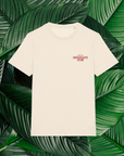 Un t-shirt beige avec un petit logo rouge sur la poitrine, comprenant le texte 'Origines Chocolate Club' dans une police stylisée. Le t-shirt est posé sur un fond composé de grandes feuilles vertes naturelles