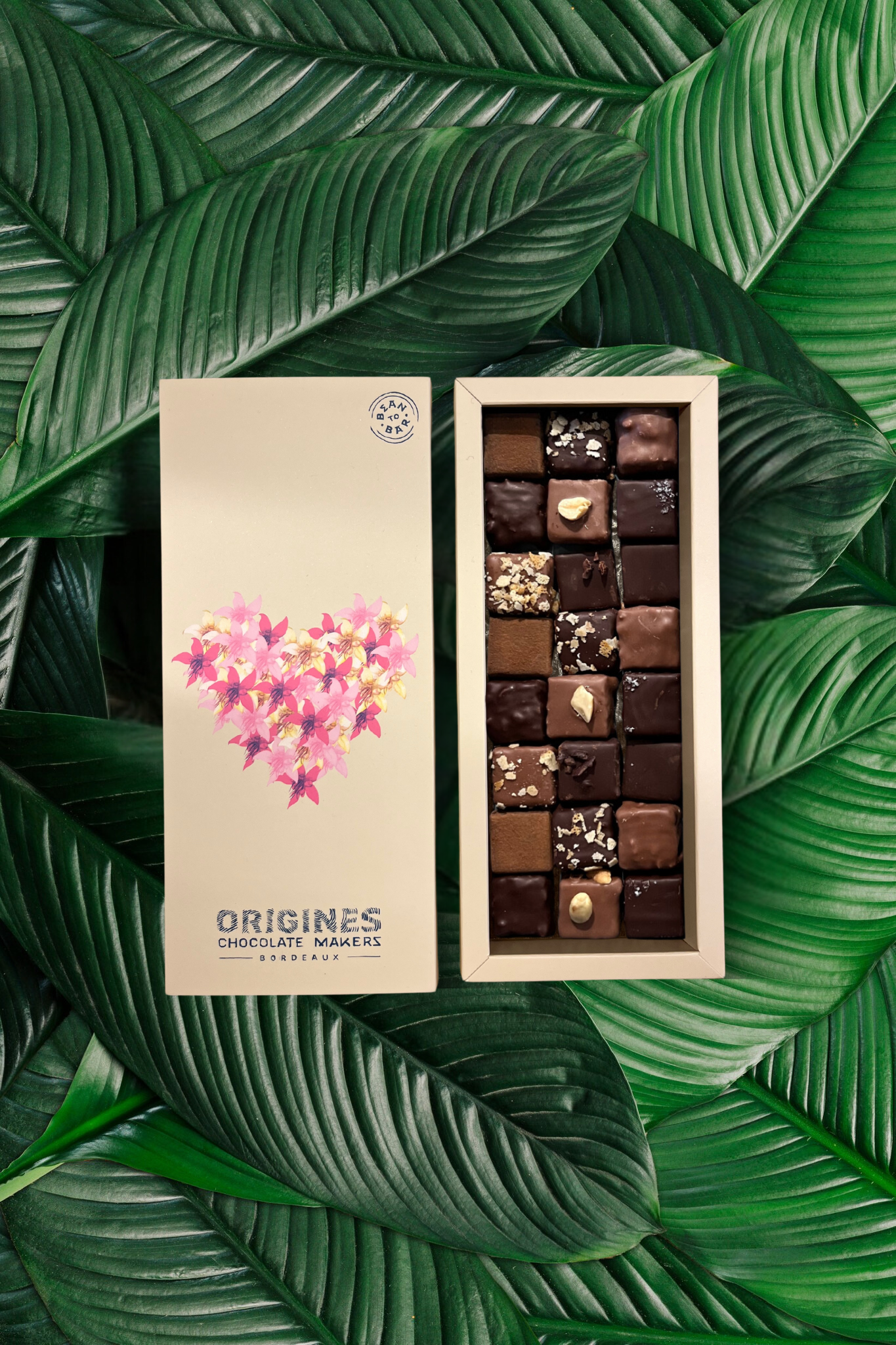 Boîte beige élégante décorée d'un cœur floral rose, signée Origines Chocolate Makers. Remplie d'un assortiment gourmand de chocolats Bean to Bar en noir et lait, posée sur un fond de feuilles tropicales.