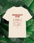 Un t-shirt beige avec un design minimaliste dessiné à la main, représentant une devanture de chocolaterie intitulée 'Origines', accompagné du texte 'Chocolate Club' en lettres stylisées. Le t-shirt est posé sur un fond composé de grandes feuilles vertes naturelles.