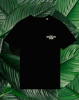 Un t-shirt noir avec un petit logo beige sur la poitrine, comprenant le texte 'Origines Chocolate Club' dans une police stylisée. Le t-shirt est posé sur un fond composé de grandes feuilles vertes naturelles