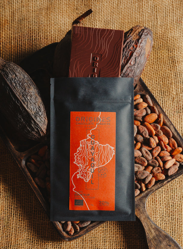 Tablette de chocolat noir Origines Chocolate Makers, emballée dans un sachet noir avec une étiquette orangeindiquant 70 % de cacao provenant d"Equateur, entourée de fèves de cacao et de cabosses sur un fond en toile de jute.