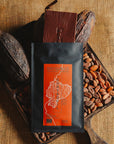 Tablette de chocolat noir Origines Chocolate Makers, emballée dans un sachet noir avec une étiquette orangeindiquant 70 % de cacao provenant d"Equateur, entourée de fèves de cacao et de cabosses sur un fond en toile de jute.