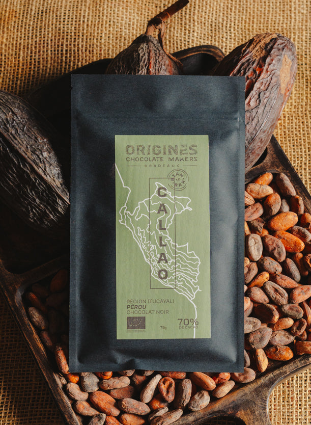 Tablette de chocolat noir Origines Chocolate Makers, emballée dans un sachet noir avec une étiquette verte indiquant 70 % de cacao provenant du Pérou, entourée de fèves de cacao et de cabosses sur un fond en toile de jute.
