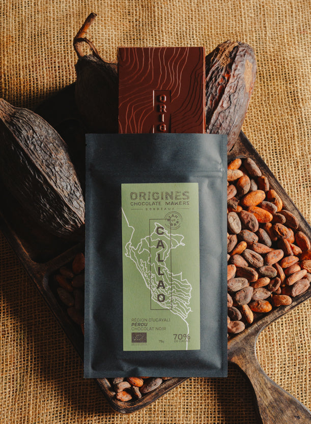 Tablette de chocolat noir Origines Chocolate Makers, emballée dans un sachet noir avec une étiquette verte indiquant 70 % de cacao provenant du Pérou, entourée de fèves de cacao et de cabosses sur un fond en toile de jute.