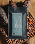 Tablette de chocolat noir Origines Chocolate Makers, emballée dans un sachet noir avec une étiquette bleue indiquant 70 % de cacao provenant de Colombie, entourée de fèves de cacao et de cabosses sur un fond en toile de jute.