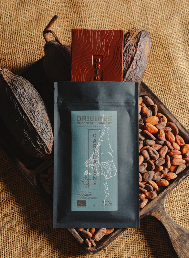 Tablette de chocolat noir Origines Chocolate Makers, emballée dans un sachet noir avec une étiquette bleue indiquant 70 % de cacao provenant de Colombie, entourée de fèves de cacao et de cabosses sur un fond en toile de jute.