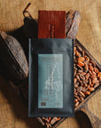 Tablette de chocolat noir Origines Chocolate Makers, emballée dans un sachet noir avec une étiquette bleue indiquant 70 % de cacao provenant de Colombie, entourée de fèves de cacao et de cabosses sur un fond en toile de jute.