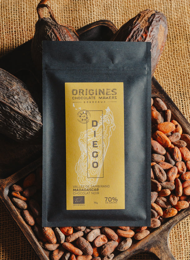Tablette de chocolat noir Origines Chocolate Makers, emballée dans un sachet noir avec une étiquette jaune indiquant 70 % de cacao provenant de Madagascar, entourée de fèves de cacao et de cabosses sur un fond en toile de jute.