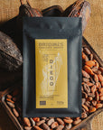 Tablette de chocolat noir Origines Chocolate Makers, emballée dans un sachet noir avec une étiquette jaune indiquant 70 % de cacao provenant de Madagascar, entourée de fèves de cacao et de cabosses sur un fond en toile de jute.