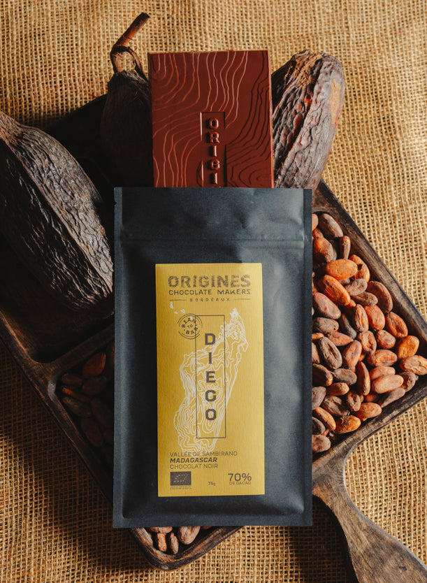Tablette de chocolat noir Origines Chocolate Makers, emballée dans un sachet noir avec une étiquette jaune indiquant 70 % de cacao provenant de Madagascar, entourée de fèves de cacao et de cabosses sur un fond en toile de jute.
