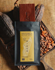Tablette de chocolat noir Origines Chocolate Makers, emballée dans un sachet noir avec une étiquette jaune indiquant 70 % de cacao provenant de Madagascar, entourée de fèves de cacao et de cabosses sur un fond en toile de jute.
