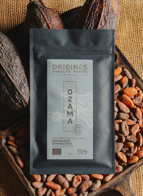 Tablette de chocolat noir Origines Chocolate Makers, emballée dans un sachet noir avec une étiquette bleue indiquant 70 % de cacao provenant de République Dominicaine, entourée de fèves de cacao et de cabosses sur un fond en toile de jute.