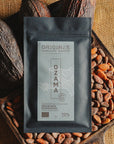 Tablette de chocolat noir Origines Chocolate Makers, emballée dans un sachet noir avec une étiquette bleue indiquant 70 % de cacao provenant de République Dominicaine, entourée de fèves de cacao et de cabosses sur un fond en toile de jute.