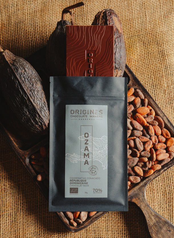 Tablette de chocolat noir Origines Chocolate Makers, emballée dans un sachet noir avec une étiquette bleue indiquant 70 % de cacao provenant de République Dominicaine, entourée de fèves de cacao et de cabosses sur un fond en toile de jute.