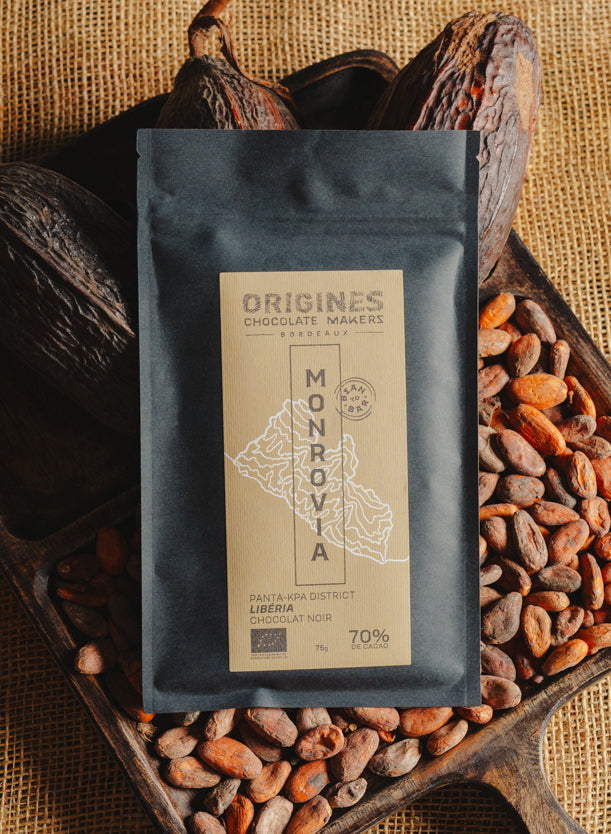 Tablette de chocolat noir Origines Chocolate Makers, emballée dans un sachet noir avec une étiquette jaune indiquant 70 % de cacao provenant du Liberia, entourée de fèves de cacao et de cabosses sur un fond en toile de jute.