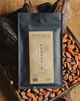 Tablette de chocolat noir Origines Chocolate Makers, emballée dans un sachet noir avec une étiquette jaune indiquant 70 % de cacao provenant du Liberia, entourée de fèves de cacao et de cabosses sur un fond en toile de jute.