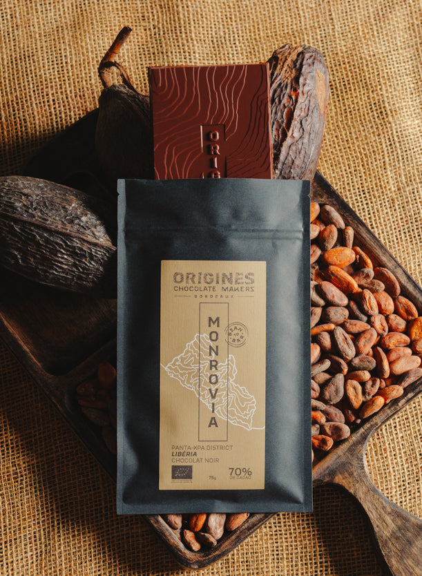 Tablette de chocolat noir Origines Chocolate Makers, emballée dans un sachet noir avec une étiquette jaune indiquant 70 % de cacao provenant du Liberia, entourée de fèves de cacao et de cabosses sur un fond en toile de jute.