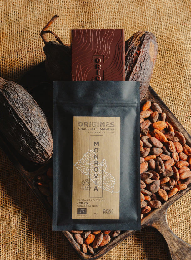 Tablette de chocolat noir Origines Chocolate Makers, emballée dans un sachet noir avec une étiquette jaune indiquant 85 % de cacao provenant du Liberia, entourée de fèves de cacao et de cabosses sur un fond en toile de jute.