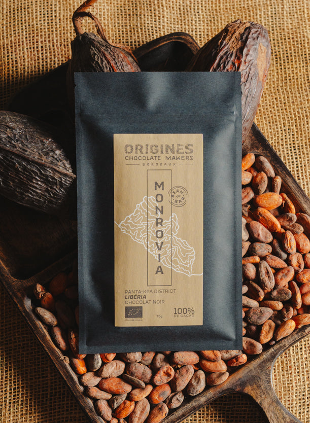 Tablette de chocolat noir Origines Chocolate Makers, emballée dans un sachet noir avec une étiquette jaune indiquant 100 % de cacao provenant du Liberia, entourée de fèves de cacao et de cabosses sur un fond en toile de jute.