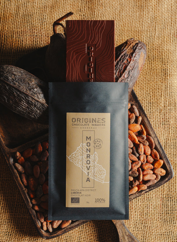 Tablette de chocolat noir Origines Chocolate Makers, emballée dans un sachet noir avec une étiquette jaune indiquant 100 % de cacao provenant du Liberia, entourée de fèves de cacao et de cabosses sur un fond en toile de jute.