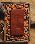 Tablette de chocolat noir au grué Origines Chocolate Makers 70 % de cacao provenant du Pérou, entourée de fèves de cacao, de cabosses et d'un bol contenant du grué de cacao sur un fond en toile de jute.