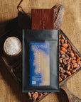 Tablette de chocolat noir à la fleur de sel du bassin d'Arcachon Origines Chocolate Makers, emballée dans un sachet noir avec une étiquette marron indiquant 70 % de cacao provenant du Pérou, entourée de fèves de cacao, de cabosses et d'un bol contenant du sel sur un fond en toile de jute.