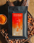 Tablette de chocolat noir au piment d'Espelette Origines Chocolate Makers, emballée dans un sachet noir avec une étiquette orange indiquant 70 % de cacao provenant du Pérou, entourée de fèves de cacao, de cabosses et d'un bol contenant du piment d'Espelette sur un fond en toile de jute.