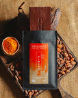 Tablette de chocolat noir au piment d'Espelette Origines Chocolate Makers, emballée dans un sachet noir avec une étiquette orange indiquant 70 % de cacao provenant du Pérou, entourée de fèves de cacao, de cabosses et d'un bol contenant du piment d'Espelette sur un fond en toile de jute.