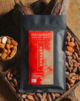 Tablette de chocolat noir au amandes de Valence Origines Chocolate Makers, emballée dans un sachet noir avec une étiquette rouge indiquant 70 % de cacao provenant du Pérou, entourée de fèves de cacao, de cabosses et d'un bol contenant des amandes sur un fond en toile de jute.