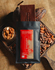 Tablette de chocolat noir au amandes de Valence Origines Chocolate Makers, emballée dans un sachet noir avec une étiquette rouge indiquant 70 % de cacao provenant du Pérou, entourée de fèves de cacao, de cabosses et d'un bol contenant des amandes sur un fond en toile de jute.