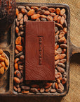 Tablette de chocolat noir au amandes de Valence Origines Chocolate Makers 70 % de cacao provenant du Pérou, entourée de fèves de cacao, de cabosses et d'un bol contenant des amandes sur un fond en toile de jute.