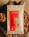 Tablette de chocolat au lait au amandes de Valence Origines Chocolate Makers, emballée dans un sachet kraft avec une étiquette rouge indiquant 38 % de cacao de São Tomé entourée de fèves de cacao, de cabosses et d'un bol contenant des amandes sur un fond en toile de jute.
