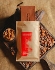 Tablette de chocolat au lait au amandes de Valence Origines Chocolate Makers, emballée dans un sachet kraft avec une étiquette rouge indiquant 38 % de cacao de São Tomé entourée de fèves de cacao, de cabosses et d'un bol contenant des amandes sur un fond en toile de jute.