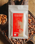 Tablette de chocolat blanc aux amandes de Valence Origines Chocolate Makers, emballée dans un sachet blanc avec une étiquette marron indiquant 35 % de cacao provenant du Pérou, entourée de fèves de cacao, de cabosses et d'un bol contenant des amandes sur un fond en toile de jute.