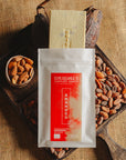 Tablette de chocolat blanc aux amandes de Valence Origines Chocolate Makers, emballée dans un sachet blanc avec une étiquette marron indiquant 35 % de cacao provenant du Pérou, entourée de fèves de cacao, de cabosses et d'un bol contenant des amandes sur un fond en toile de jute.