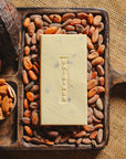 Tablette de chocolat blanc aux amandes de Valence Origines Chocolate Makers 35 % de cacao provenant du Pérou, entourée de fèves de cacao, de cabosses et d'un bol contenant des amandes sur un fond en toile de jute.