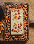 Dos d'une tablette de chocolat blanc aux amandes de Valence Origines Chocolate Makers 35 % de cacao provenant du Pérou, entourée de fèves de cacao, de cabosses et d'un bol contenant des amandes sur un fond en toile de jute.