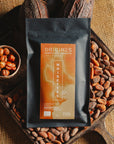 Tablette de chocolat noir au noisettes de lot et garonne Origines Chocolate Makers, emballée dans un sachet noir avec une étiquette marron indiquant 70 % de cacao provenant du Pérou, entourée de fèves de cacao, de cabosses et d'un bol contenant des noisettes sur un fond en toile de jute.