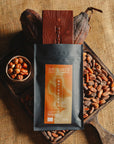 Tablette de chocolat noir au noisettes de lot et garonne Origines Chocolate Makers, emballée dans un sachet noir avec une étiquette marron indiquant 70 % de cacao provenant du Pérou, entourée de fèves de cacao, de cabosses et d'un bol contenant des noisettes sur un fond en toile de jute.