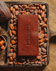 Tablette de chocolat noir au noisettes de lot et garonne Origines Chocolate Makers 70 % de cacao provenant du Pérou, entourée de fèves de cacao, de cabosses et d'un bol contenant des noisettes sur un fond en toile de jute.
