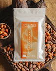 Tablette de chocolat blanc au noisettes de lot et garonne Origines Chocolate Makers, emballée dans un sachet blanc avec une étiquette marron indiquant 35 % de cacao, entourée de fèves de cacao, de cabosses et d'un bol contenant des noisettes sur un fond en toile de jute.