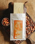 Tablette de chocolat blanc au noisettes de lot et garonne Origines Chocolate Makers, emballée dans un sachet blanc avec une étiquette marron indiquant 35 % de cacao entourée de fèves de cacao, de cabosses et d'un bol contenant des noisettes sur un fond en toile de jute.