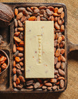 Tablette de chocolat blanc au noisettes de lot et garonne Origines Chocolate Makers 35 % de cacao, entourée de fèves de cacao, de cabosses et d'un bol contenant des noisettes sur un fond en toile de jute.