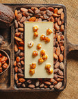 Dos d'une tablette de chocolat blanc au noisettes de lot et garonne Origines Chocolate Makers 35 % de cacao, entourée de fèves de cacao, de cabosses et d'un bol contenant des noisettes sur un fond en toile de jute.