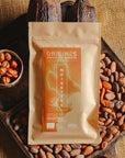 Tablette de chocolat au lait au noisettes de lot et garonne Origines Chocolate Makers, emballée dans un sachet kraft avec une étiquette marron indiquant 38 % de cacao de São Tomé entourée de fèves de cacao, de cabosses et d'un bol contenant des noisettes sur un fond en toile de jute.